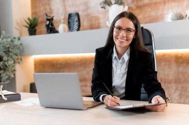 Başarılı, genç bir iş kadınının portresi, broker, finansal danışman, satış müdürü, modern bir ofiste, şık iş elbiseleriyle oturuyor, gazetelerle çalışıyor, kameraya bakıyor, gülümsüyor.