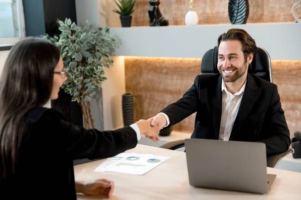 Successful young adult caucasian business colleagues, entrepreneurs, directors, sit at a desktop in modern office, made a good deal, agreed of cooperation, shake hands to each other, smiling — Stock Photo, Image