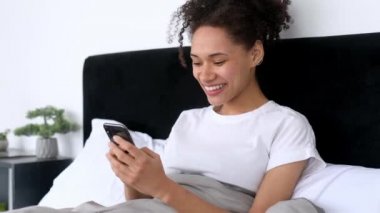 Mutlu Afro-Amerikan güzel kızı akıllı telefonunu kullanıyor, sabahları yatakta yatarken, internette ve sosyal ağlarda gezinirken, arkadaşlarıyla ya da ailesiyle mesajlaşırken, iyi haberleri öğreniyor, gülümsüyor.