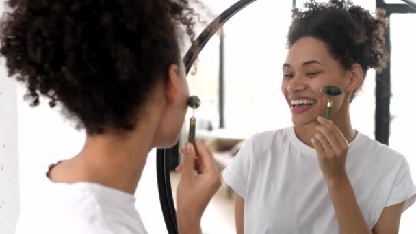 Cuidados com a pele. Encantadora bela jovem afro-americana usando rolo derma para massagear a pele facial, em casa na frente de um espelho, torna a pele flexível, evita rugas, sorrisos — Vídeo de Stock