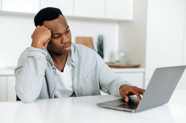 Bored frustrated african american guy, freelancer, designer, sitting at a table at home, dressed in casual clothes, working at a laptop, tired, in needs of rest or break, upset