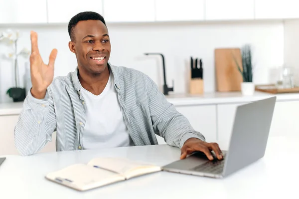 성공적 인 현대 아프리카 계 미국인 남성, 프리랜서, 디자이너 또는 IT 전문가는 노트북을 사용하여 동료나 고객 과 대화하고 우호적 인 미소를 지으며 집에서 일한다. 멀리 떨어져 있는 의사소통 — 스톡 사진