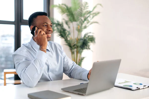 Bem-sucedido confiante ocupado afro-americano homem de negócios, agente imobiliário, gerente, sentado em um escritório moderno, falando ao telefone com colegas ou clientes, ter uma conversa importante, sorrindo — Fotografia de Stock