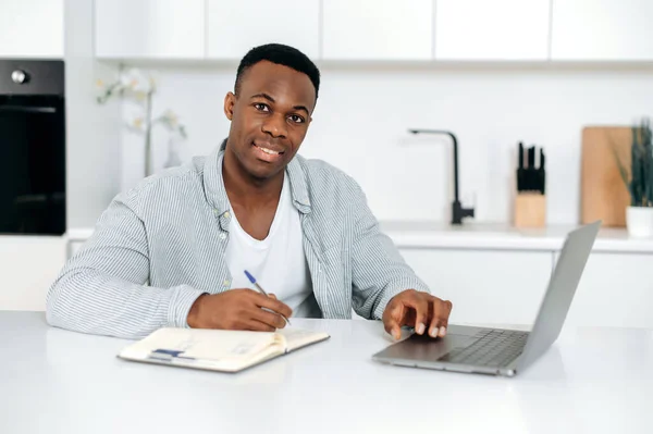 Atractivo chico afroamericano, estudiante, freelancer, se sienta en una mesa en la cocina, utiliza computadora portátil y bloc de notas para el aprendizaje en línea, mira a la cámara, escucha una conferencia, toma notas, sonríe amigable — Foto de Stock