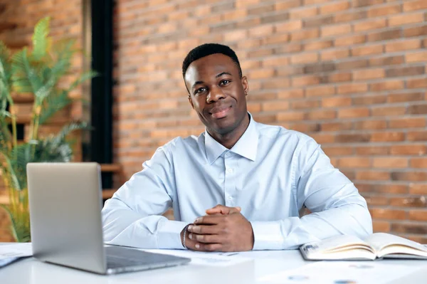 Jovem afro-americano agradável a trabalhar num computador num escritório moderno. Retrato de um empresário de sucesso, freelancer, gerente, vestindo uma camisa formal, olhando para a câmera, sorrindo amigável. — Fotografia de Stock