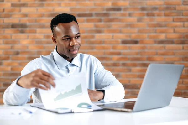 Empresario afroamericano enfocado analizando gráficos financieros, ganancias, sentado en el lugar de trabajo. Estudiante negro inteligente estudia en línea, escuchar conferencias en línea, freelancer es upskills, usando el ordenador portátil — Foto de Stock