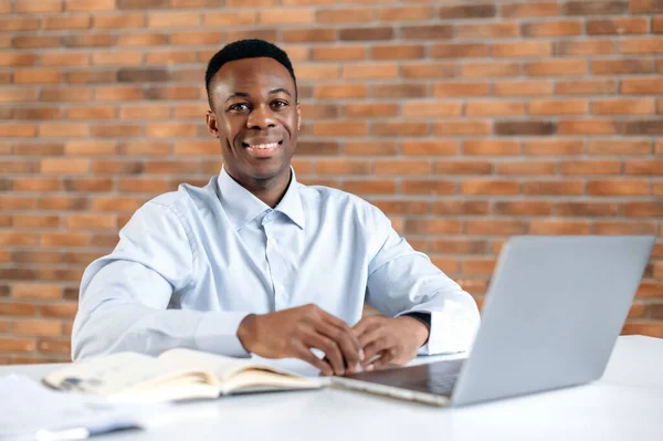 현대 사무실의 매력적 인 아프리카 계 미국인 남자의 초상화. 성공 한 똑똑 한 사람, 회사의 최고 경영자나 트레이더, 직장에서 노트북을 가지고 일하는 사람, 카메라를 보고 웃으세요. — 스톡 사진