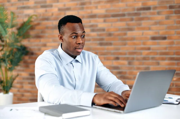 Chocado surpreso afro-americano homem de negócios, secretário, trabalhador de escritório, sentado em sua mesa no escritório, olhando para laptop em espanto, recebendo notícias inesperadas, ganhar ou fazer grande lucro — Fotografia de Stock