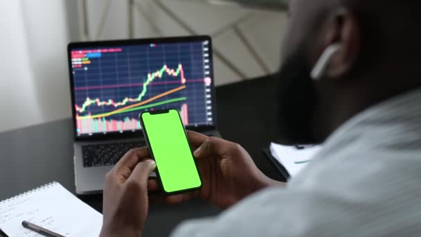 Afrykański inwestor kryptowaluta handlowiec posiada w ręku telefon komórkowy z zielonym ekranem makiety, analizować dynamikę rynku akcji, studiuje wykresy, wykorzystuje aplikację mobilną handlu aplikacji, w biurze — Wideo stockowe