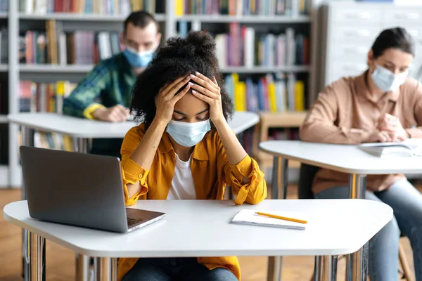 Educação durante a pandemia. Frustrada, trabalhada demais menina estudante afro-americana vestindo máscara médica protetora senta-se em uma mesa na biblioteca, se preparando para exames, cansado, colegas de classe sentar no fundo — Fotografia de Stock