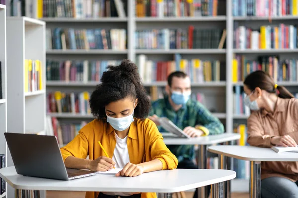 Estudantes durante pandemia com máscaras médicas. Concentrada menina americana africana usa máscara médica protetora sentar à mesa na biblioteca, se preparando para o exame, toma notas, colegas de classe sentar no fundo — Fotografia de Stock