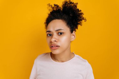 Şaşkın, heyecanlı, şaşkın, kıvırcık saçlı Afro-Amerikan genç kadın kameraya sorguluyor, anlayamıyor, pembe arka planda duruyor.