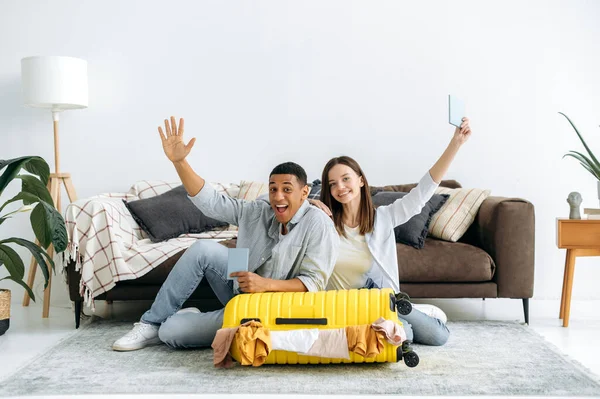 Farklı milletlerden gelen mutlu bir çift, oturma odasında yerde oturuyor, bavullarını topluyor, bir yolculuğa hazırlanıyor, pasaportlarını tutuyor, kameraya neşeyle gülümsüyor.