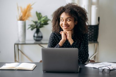 Tatmin olmuş, koyu tenli, kıvırcık saçlı genç kadın, ofis çalışanı, yönetici ya da finansör, modern ofiste bir masada oturuyor, işten dinleniyor, ara veriyor, uzaklara bakıyor, gülümsüyor, dinlenmeyi hayal ediyor.