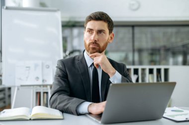 Düşünceli, zeki, beyaz erkek girişimci, finans müdürü, resmi takım elbiseli CEO, modern ofisin masasında oturuyor, başka yere bakıyor, yeni projeyi, iş stratejisini, pazarlama fikirlerini düşünüyor.