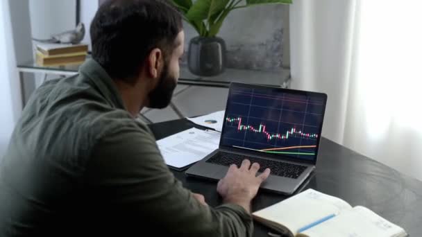 Online kereskedelem. Sikeres indiai vagy arab kereskedő, aki laptoppal dolgozik, miközben az irodájában ül, elemzi a kereskedési táblázatokat, megjósolja a kriptovaluta emelkedését vagy bukását, jegyzetel. — Stock videók