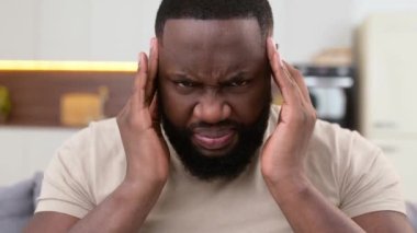 Afro-Amerikan sağlıksız bir adam evde oturma odasında oturuyor, baş ağrısı çekiyor, migren oluyor, şakaklarına masaj yapıyor, dinlenmeye, uyumaya, tedaviye ihtiyacı var. Migren konsepti
