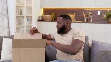 Heyecanlı bir Afrikalı Amerikalı mutlu adam, evde bir paket aldı, bir karton kutu açtı, aldığı kıyafetleri inceledi, yeni bir alışverişe sevindi. İnternetten alışveriş, ev teslimatı konsepti