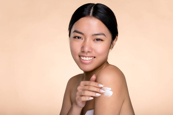 Skin body care concept. Beautiful asian woman gently applying body lotion, cream on her naked shoulder, prevents skin aging, moisturizes, standing on isolated beige background, look at camera, smiles