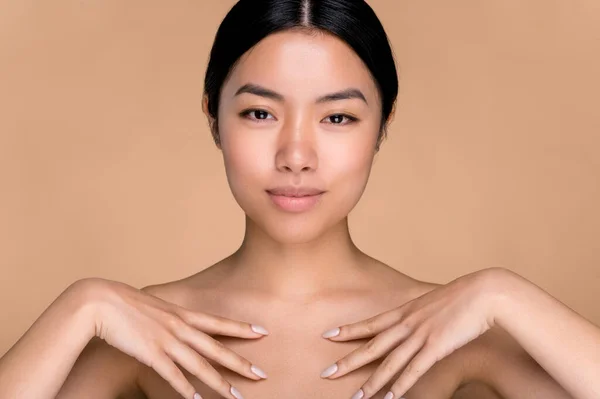 Beauty, care skin concept. Beautiful chinese brunette young woman, with perfect skin and naked shoulders, posing with hands near collarbone at isolated beige background, look at camera, smile pleasant