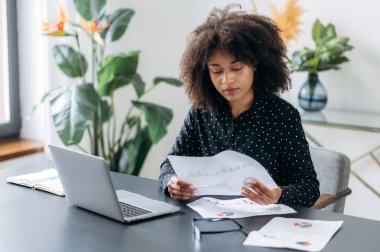 Odaklanmış koyu tenli kadın, serbest çalışan, yönetici, şirket müdürü, bir dizüstü bilgisayarın başında oturuyor, belgelerle çalışıyor, dinamikleri analiz ediyor, çalışma stratejisi geliştiriyor, iş planı yapıyor.