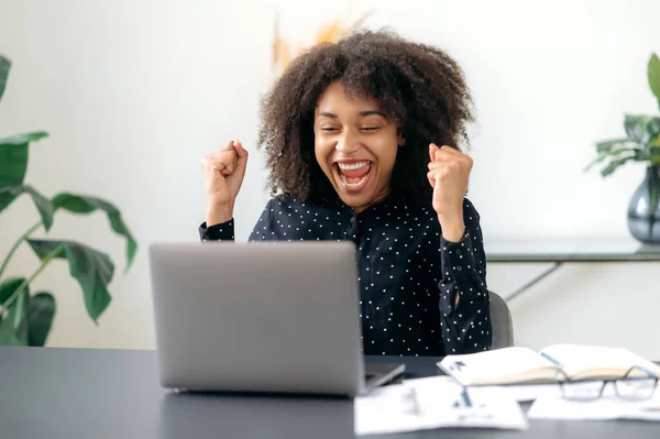 Terkejut dengan kesuksesan wanita bisnis Amerika yang percaya diri, pialang, manajer, duduk di meja dengan laptop, menikmati keuntungan besar, kemenangan, kabar baik, isyarat dengan kepalan tangan, tersenyum — Stok Foto