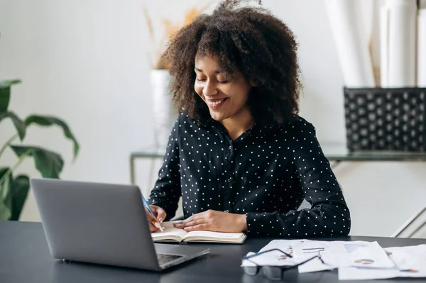 Directeur d'entreprise afro-américain ou pigiste travaillant dans le bureau. Belle femme aux cheveux bouclés intelligente et confiante, assise devant un ordinateur portable, prenant des notes dans un carnet, écoutant un webinaire, souriant — Photo