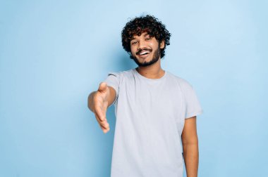 Genç, pozitif kıvırcık saçlı, sıradan tişörtler giyen Arap ya da Hint vatandaşı. Dostça bir selamlama, anlaşma ve hoş geldin olarak el sıkışma sunuyor. 