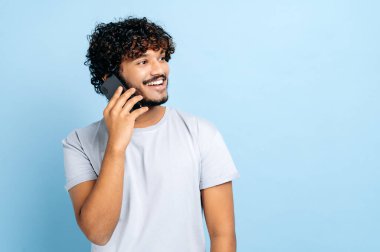 Cep telefonu sohbet konsepti. Tatmin olmuş Arap ya da Hintli çekici adam, akıllı telefonunu kullanıyor, cep telefonuyla konuşuyor, izole edilmiş mavi arka planda duruyor, başka tarafa bakıyor, güler yüzlü dost canlısı.