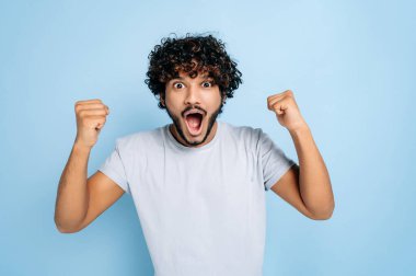 Hevesli, şaşkın Hintli ya da Arap tişörtlü adam, başarı, zafer, kazanma, yumruklarla hareket etme, kameraya bakma, gülümseme, izole edilmiş mavi bir arka planda durma sevinci içinde.