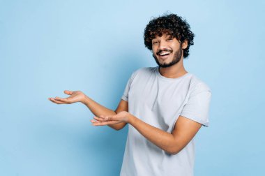 Pozitif çekici kıvırcık saçlı yerli ya da Arap tişörtlü adam kameraya bakıyor ve elleri boş alanda, izole edilmiş mavi arka planda duruyor, gülümsüyor. Model konsepti