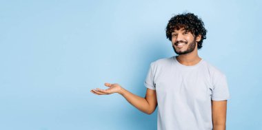 Dost canlısı Hintli ya da Arap bir adamın günlük tişörtlü panoramik fotoğrafı, kameraya bakıyor ve boş alanda el ele gösteriyor, izole edilmiş mavi arka planda duruyor, gülümsüyor. Model konsepti