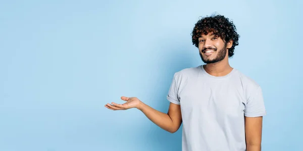 Panoramic photo of friendly indian or arabian guy in casual t-shirt, looks at the camera and points hand to the side, at empty space, stands on isolated blue background, smiling. Mock-up concept — Stock Photo, Image