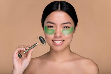 Güzellik prosedürleri. Gözlerinin altında kozmetik kolajen yeşil bantları olan, mükemmel bakımlı ciltli Çinli kadın, yeşim taşlı yüz masajı yapan, izole edilmiş bej arka planda duruyor, gülümsüyor.