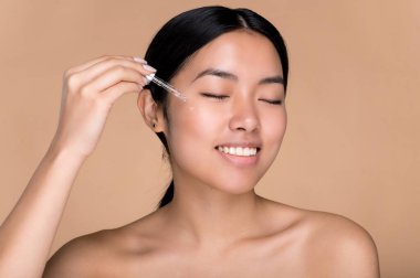 Güzellik, cilt bakımı konsepti. Mükemmel ciltli, çıplak omuzlu, ciltli, cilt bakımlı, yüz yağlı, nemlendirici hiyalüronik serum kullanan, gözleri kapalı, izole edilmiş bej arka planı olan güzel Çinli genç bir kadın.