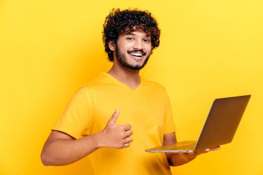 Kıvırcık saçlı, tişörtlü, elinde dizüstü bilgisayarla kameraya gülümseyen, işaret eden, izole edilmiş turuncu arka planda duran mutlu Arap ya da Hintli adam.