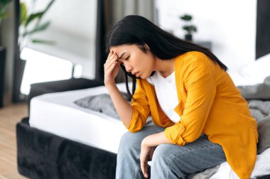 Üzgün Çinli esmer kadın, gündelik giysiler içinde, evde yatakta oturuyor, kişisel problemler, haberler, yalnızlık, stres, belirsizlik yüzünden üzgün.
