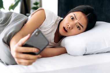 Şaşırmış Asyalı bir kız sabah yatakta yatarken, akıllı telefonuna korkmuş görünüyor, çalar saati fazla kaçırmış, duygusal yüz ifadesi, olumsuz duygu, beklenmedik haberler.