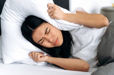 Asyalı genç bir kadın, evde yatarken, kulaklarını yastıkla kapatıyor, uyumaya çalışıyor, gürültüden kendini kapatıyor, kötü bir rüya görüyor, huzursuz bir rüya. Uyku ve dinlenme konsepti