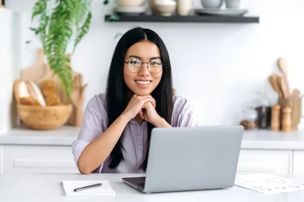 Hezká, pozitivní chytrá asijská brunetka s brýlemi, designérka, nezávislá nebo studentka, sedí doma v kuchyni s laptopem pro vzdálenou práci nebo studium, dívá se do kamery, přátelsky se usmívá — Stock fotografie