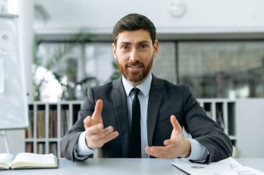 Başarılı bir beyaz adam, girişimci, avukat, yönetici modern bir ofiste şık iş elbiseleriyle oturuyor, iş arkadaşlarıyla çevrimiçi video toplantısı yapıyor, konuşuyor, el kol hareketi yapıyor.
