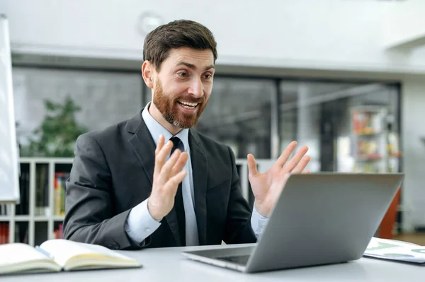 Surpreso, homem de negócios caucasiano, diretor da empresa, programador, senta-se em uma área de trabalho na frente de uma tela de laptop no escritório, conversa com um colega em uma chamada de vídeo, descobre boas notícias sobre um bom negócio — Fotografia de Stock