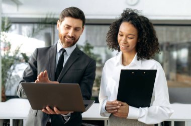 Pozitif dost canlısı iş arkadaşları, esmer tenli güzel bir kadın ve nüfuzlu beyaz bir adam, modern bir ofiste dizüstü bilgisayardan bir rapor izliyor, iş planı ve stratejisi geliştiriyor, gülümsüyor.