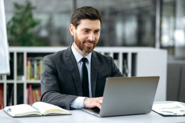 Yakışıklı beyaz adam, menajer, avukat, ekonomist, bir dizüstü bilgisayarın önünde, modern bir ofiste, şık bir iş elbisesi içinde, müşterisi veya çalışanı ile iş yazışmaları yapıyor, gülümsüyor.
