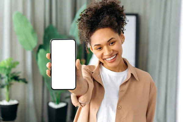 Afroamerikanerin mit lockigem Haar in Freizeitkleidung, zu Hause stehend, Smartphone in der Hand, weißer Bildschirm für Werbung, in die Kamera guckend, glücklich lächelnd — Stockfoto