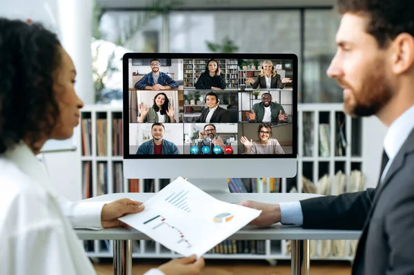 An international group of business partners gathered for an online video meeting. Business colleagues, a man and a woman sit in office in front of a computer screen, study business papers together — Stock Photo, Image