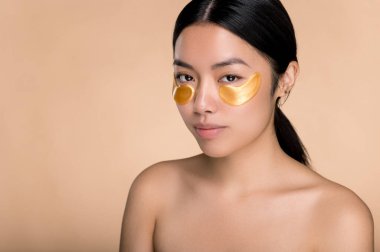 Yüz prosedürlerini önemser. Mükemmel cilde sahip Asyalı güzel bir kız, gözlerinin altına kozmetik altın bantlar takar, göz çevresindeki deriyi önemser, kırışıklıkları önler, kameraya bakar, izole edilmiş bej arka plan