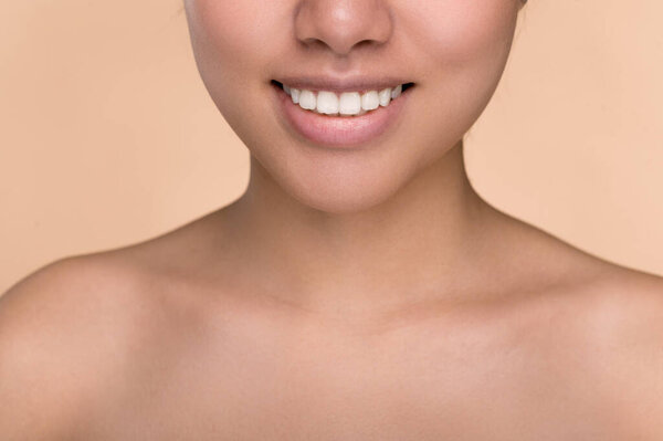 Half face of Asian girl. Chinese young woman with perfect skin demonstrates her smile, perfect white well-groomed teeth, stands on an isolated beige background with naked shoulders. Dentistry concept