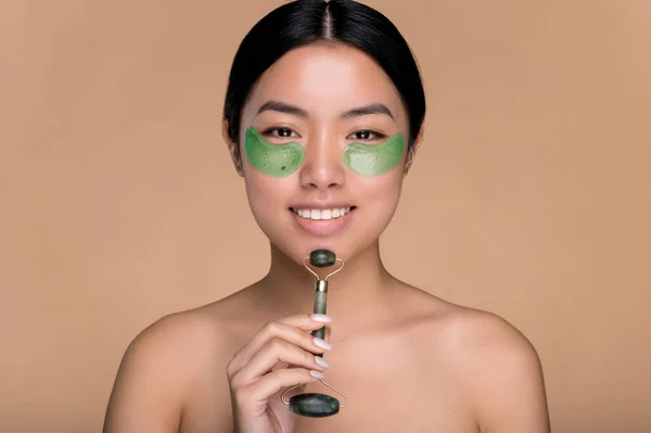 Prosedur kecantikan kosmetik. Gadis Cina dengan kulit rapi, dengan plester kolagen hijau kosmetik di bawah mata, memijat kulit wajah dengan penggulung giok, berdiri di latar belakang krem yang terisolasi, tersenyum — Stok Foto