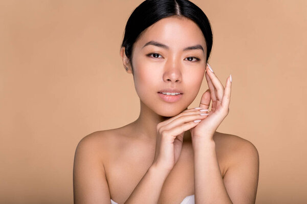 Beauty, care and youth concept. Lovely japanese brunette young woman, with perfect skin and naked shoulders, posing with hands near face at isolated beige background, looking at camera, smiles